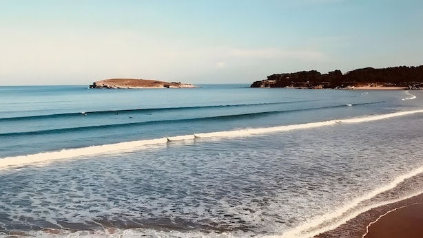 Ein Strand mit Wellen, einem sandigen Ufer und einem Felsen im Hintergrund.