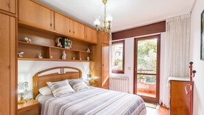Ein Schlafzimmer mit Holz-Kopfteil, einem Bett mit gestreifter Bettwäsche, einem Nachttisch mit Lampe, einer hölzernen Kommode und Blick durch eine offene Tür auf einen Außenbereich.