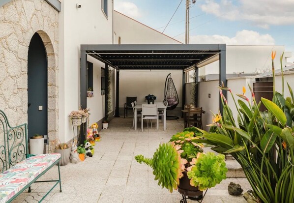 Un patio couvert avec une table et des chaises, entouré de plantes en pot et agrémenté d’un fauteuil suspendu.