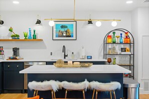 A modern kitchen with a central island, bar stools, a coffee machine, and a decorative shelf with colorful vases.