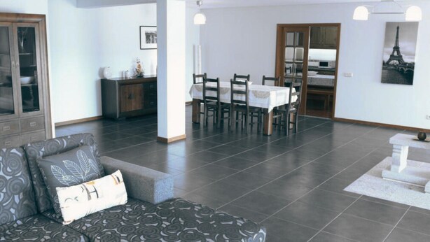 Un vaste espace de vie comprenant un canapé à motifs foncés, une table à manger avec des chaises, et une cuisine visible à travers une porte ouverte.