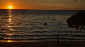 Ein Sonnenuntergang über dem Meer mit einem Strand und einem Gebäude mit einem Reetdach.