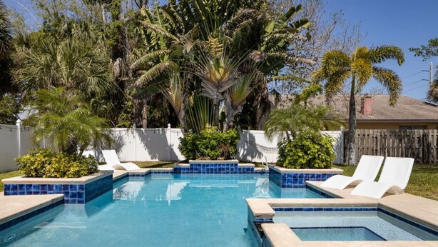 A rectangular swimming pool with blue tiles, surrounded by white lounge chairs and palm trees. - Cozy Cottage, 2 Queens, 1 Twin, Patio, Shared Pool (Melbourne)