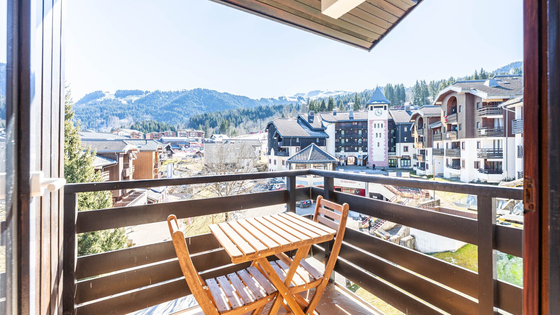 Ein Balkon mit Holztisch und Stuhl, mit Blick auf eine Stadt, mit Gebäuden und Bergen im Hintergrund.