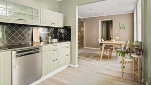 A modern kitchen with a dishwasher, a dining area with a wooden table and chairs, and a view into a living room with a bookshelf.