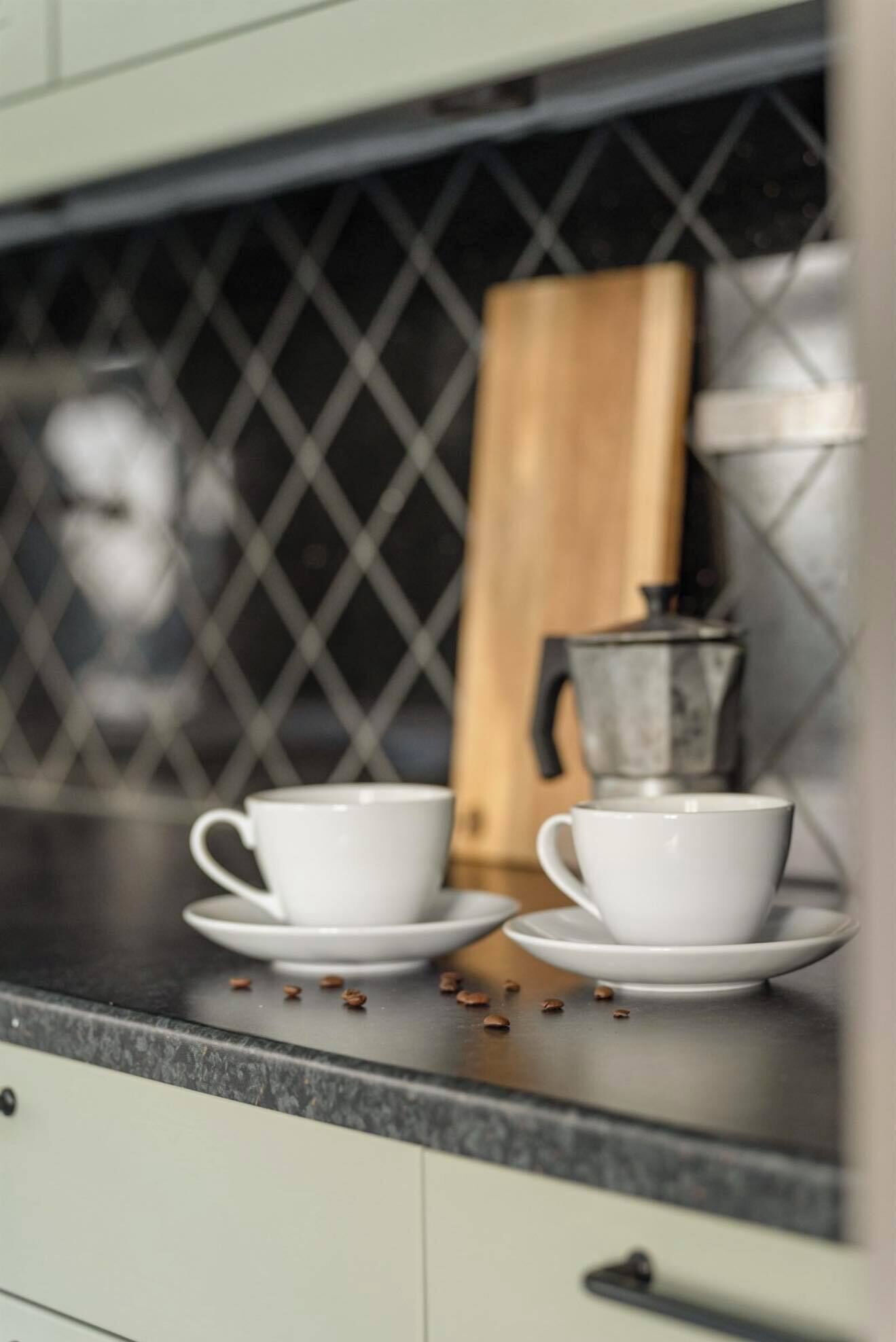 A kitchen counter with two white cups and saucers, a coffee maker, and scattered coffee beans.