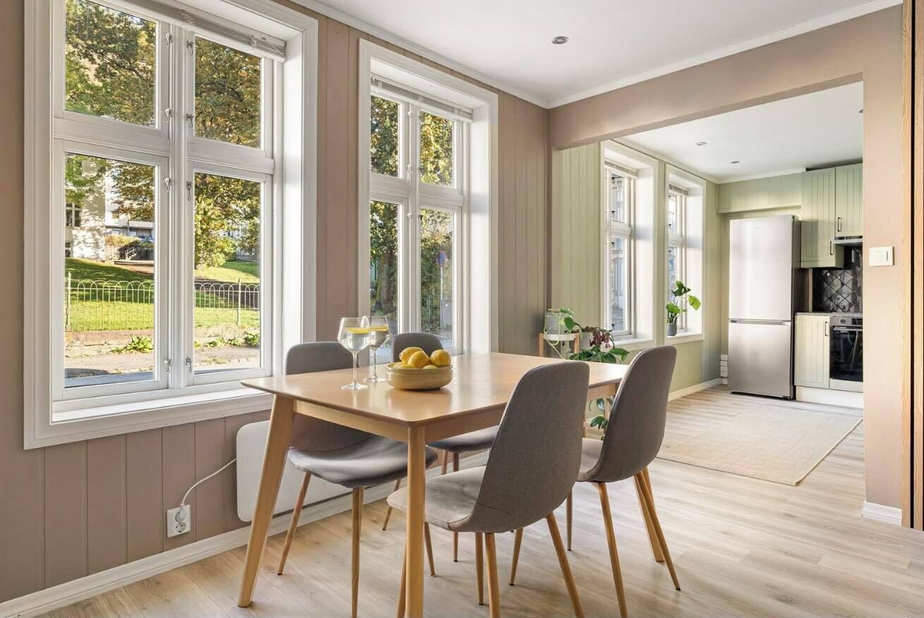Een eetgedeelte met een houten tafel en vier stoelen, grote ramen met uitzicht op groen, en een keuken op de achtergrond.