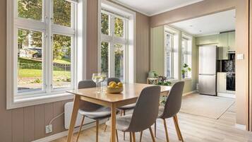 Een eetgedeelte met een houten tafel en vier stoelen, grote ramen met uitzicht op groen, en een keuken op de achtergrond.