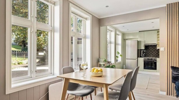A dining area with a table and chairs, large windows, a kitchen in the background, and a view of the outdoors.
