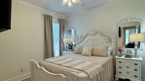 Ein Schlafzimmer mit einem Bett, einem Nachttisch, einem Spiegel, einem Deckenventilator und einem Fenster mit Vorhängen.