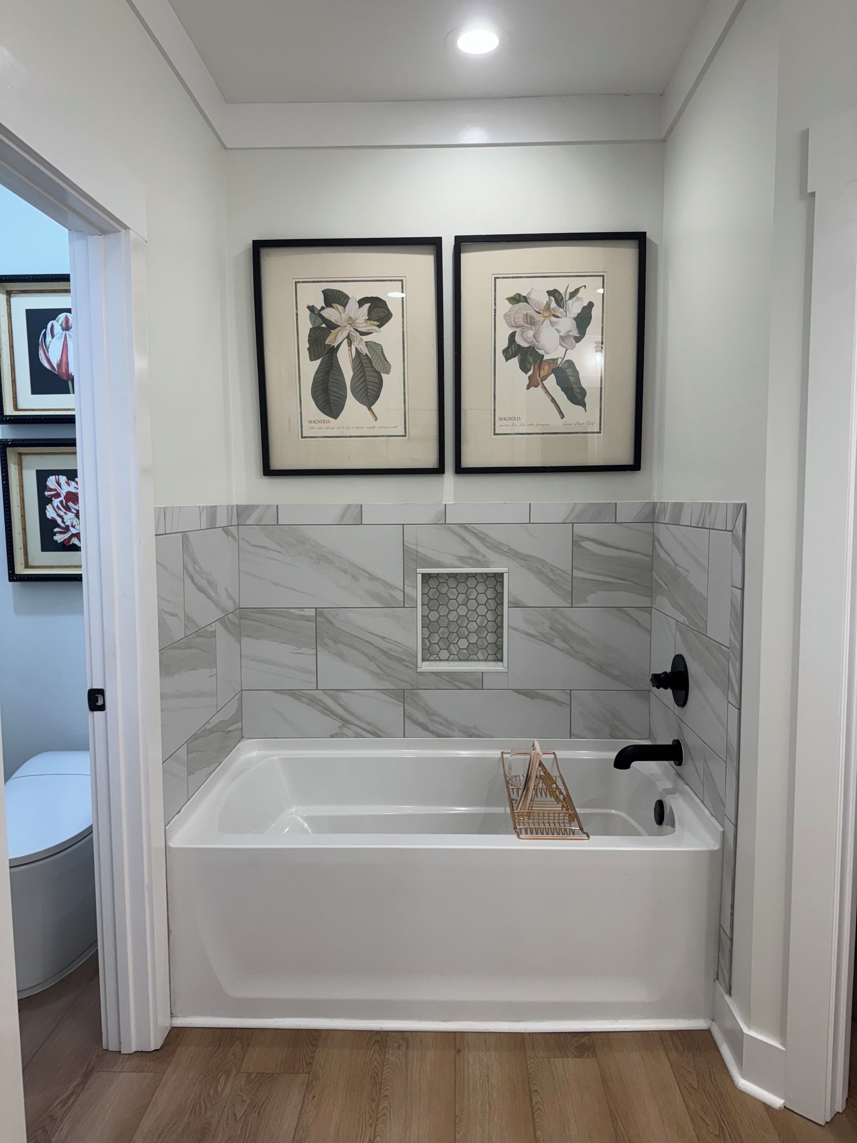 Une salle de bain moderne avec une baignoire blanche, des carreaux de marbre et deux reproductions botaniques encadrées accrochées au mur.