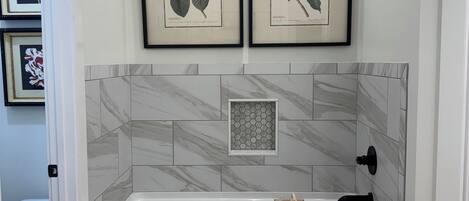 Une salle de bain moderne avec une baignoire blanche, des carreaux de marbre et deux reproductions botaniques encadrées accrochées au mur.