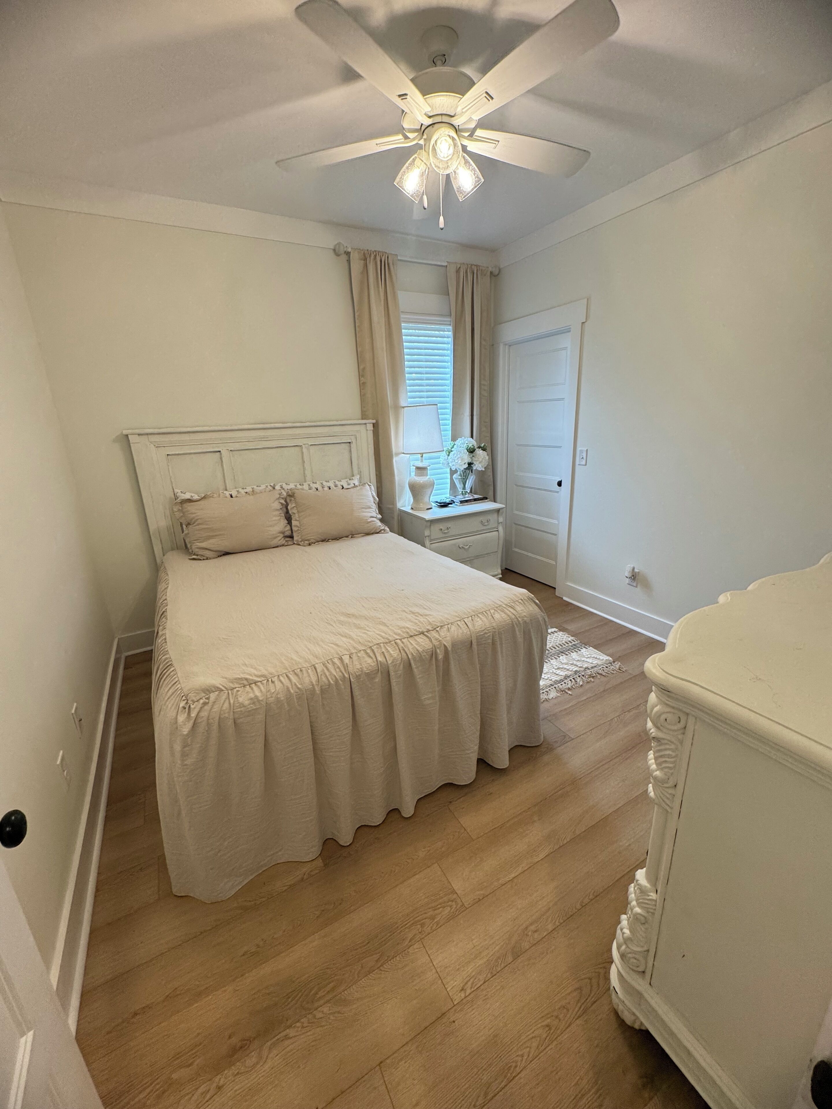 A bedroom with a bed, a nightstand, a dresser, a ceiling fan, and a window with curtains.
