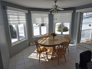 A dining area with a round table, six wooden chairs, a ceiling fan, and large windows with blinds.