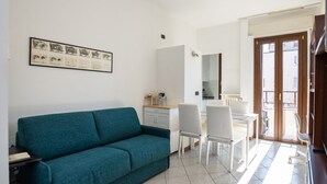 Una sala de estar moderna con un sofá color turquesa, un comedor con sillas blancas y una cocina visible a través de un umbral abierto.
