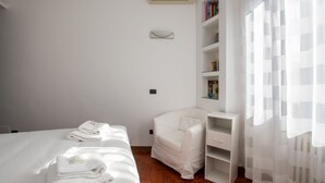 Una habitación de hotel moderna con una cama bien hecha, un sillón blanco, una mesita de noche pequeña y una estantería.