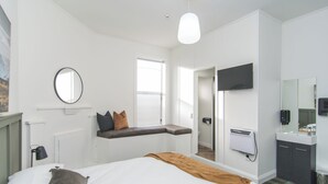Una habitación de hotel moderna con una cama grande, un banco junto a la ventana, un televisor de pantalla plana, un espejo y un baño con lavamanos e inodoro.