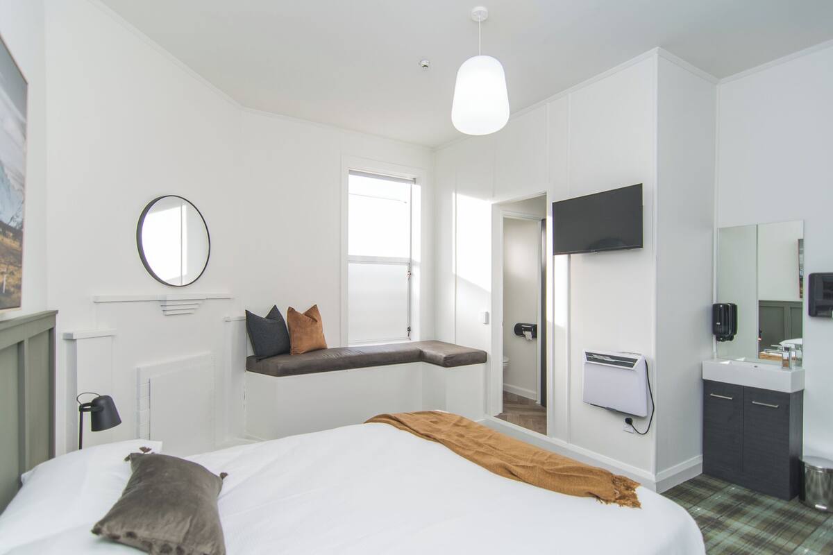 A modern hotel room with a large bed, a bench by the window, a flat-screen TV, a mirror, and a bathroom with a sink and toilet.