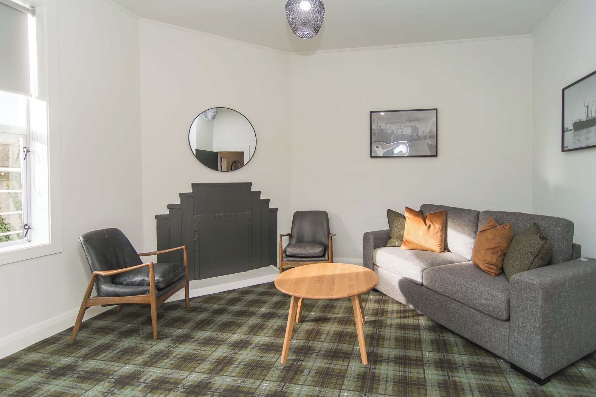 A living room with a grey sofa, two armchairs, a round wooden coffee table, a fireplace, a mirror, and two framed pictures on the wall.