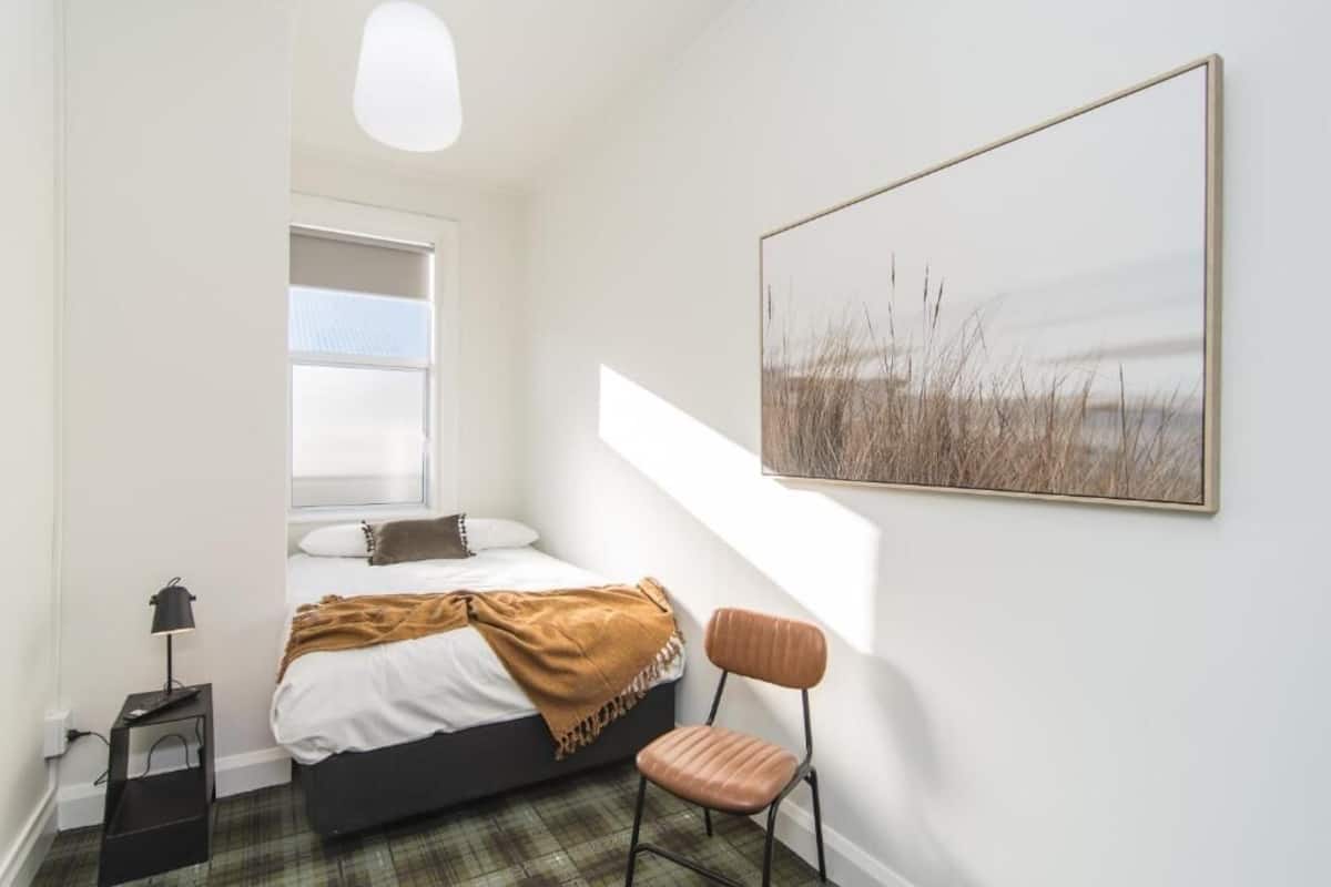 A bedroom with a bed, a chair, a bedside table, a lamp, and a large window.