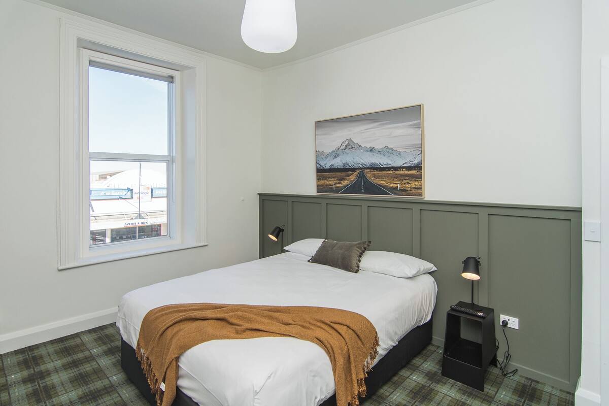 A bedroom with a bed, a window, a painting, and a nightstand.
