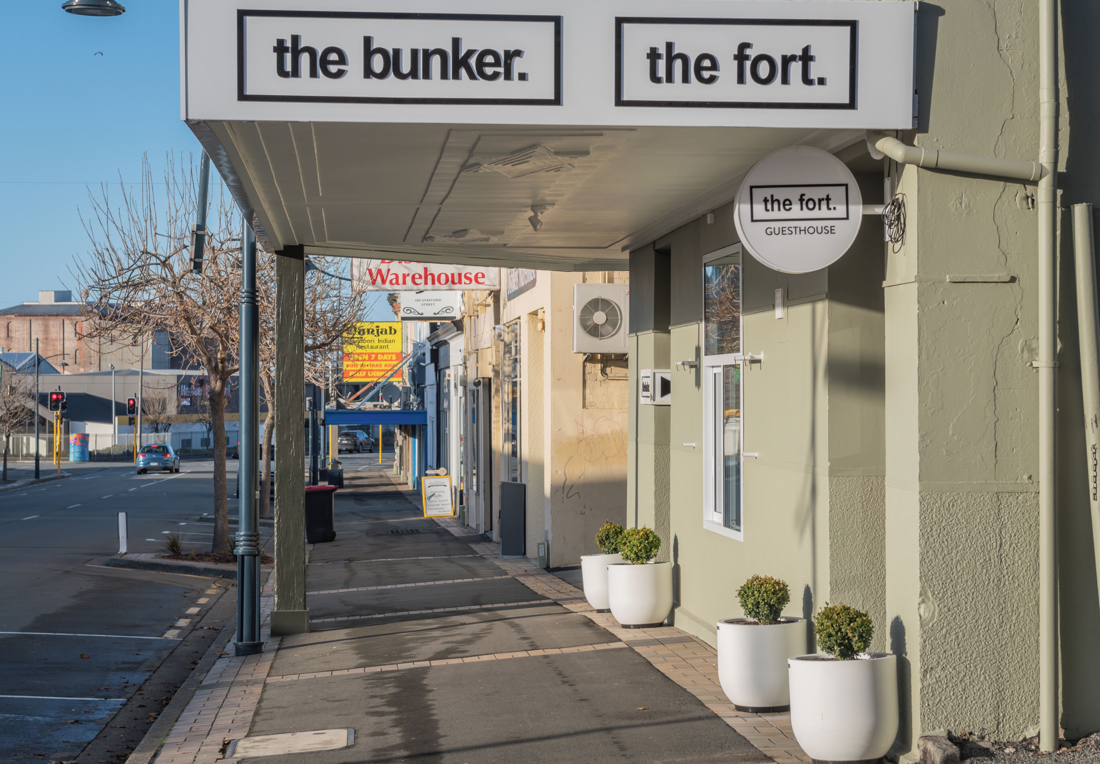 Un edificio con letreros que indican 'the bunker' y 'the fort', indicando casas de huéspedes. Hay plantas en macetas en la acera, y se ve un letrero de 'Warehouse' al fondo.