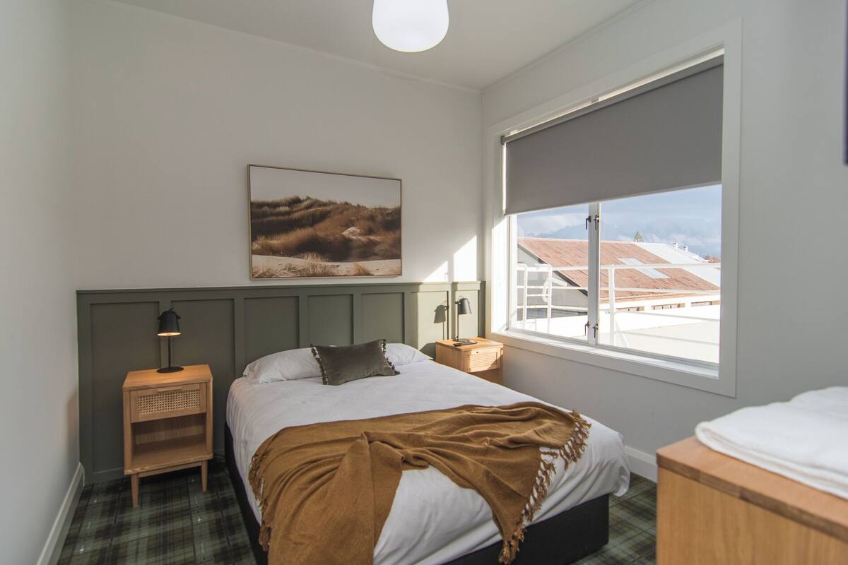 A bedroom with a bed, a nightstand, a lamp, a window with a view of buildings, and a painting on the wall.