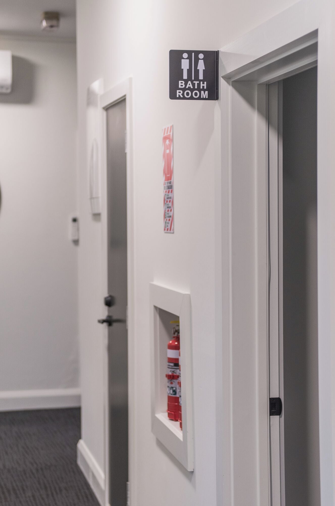Un couloir avec une pancarte indiquant l’emplacement des toilettes, un extincteur fixé au mur et une porte.