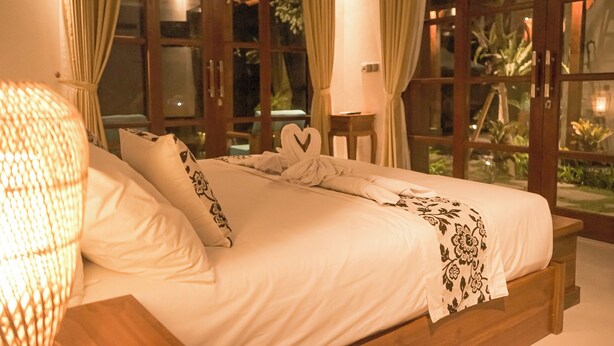 A neatly made bed with white linens and decorative pillows, a bedside table with a lamp, and large windows with curtains. - Villa Puri Kembar – A Serene Escape with a Private Pool & Stunning Views (Bali)