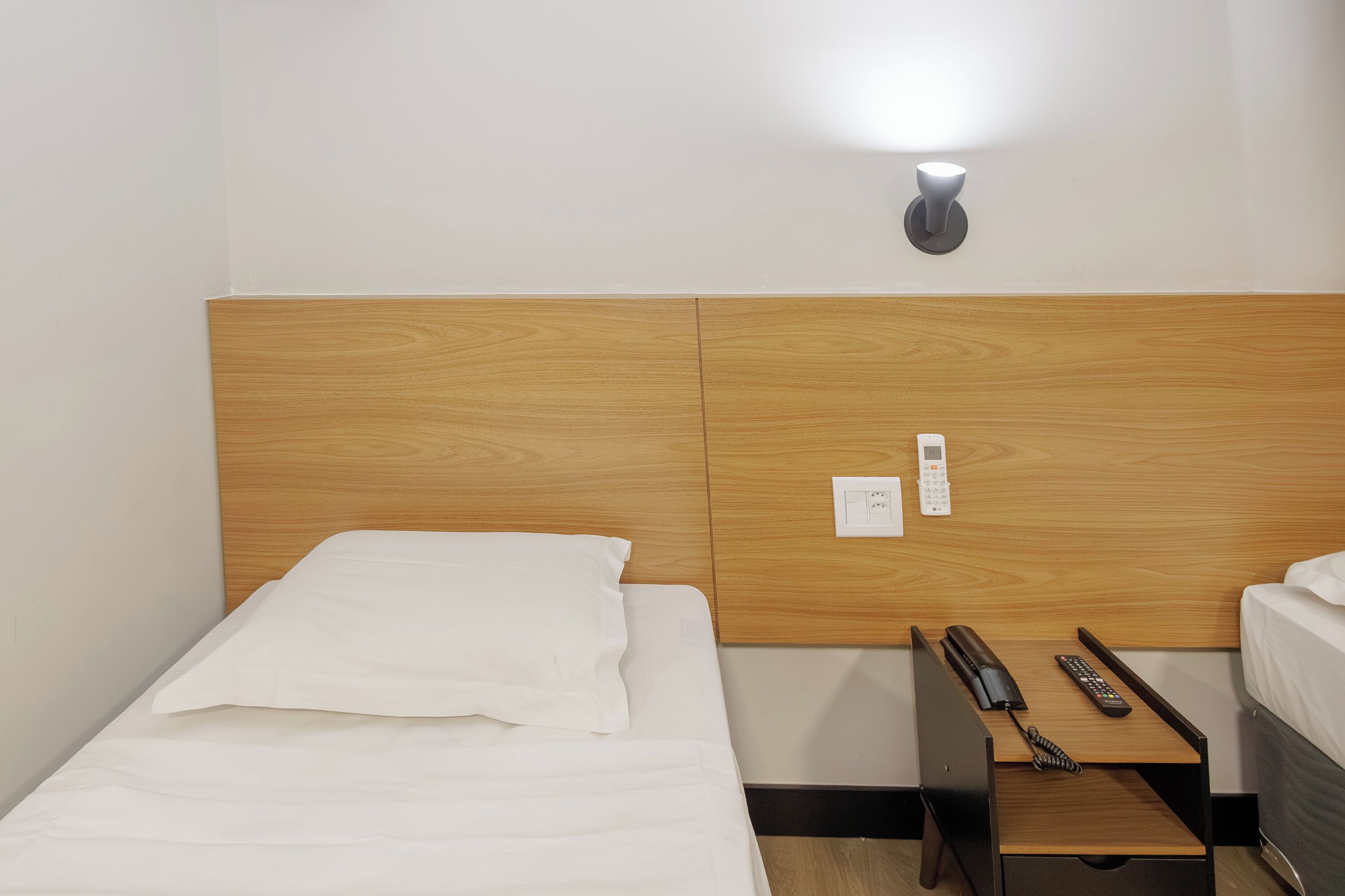 A hotel room with a wooden headboard, a bedside table with a phone and remote controls, and a wall-mounted light.