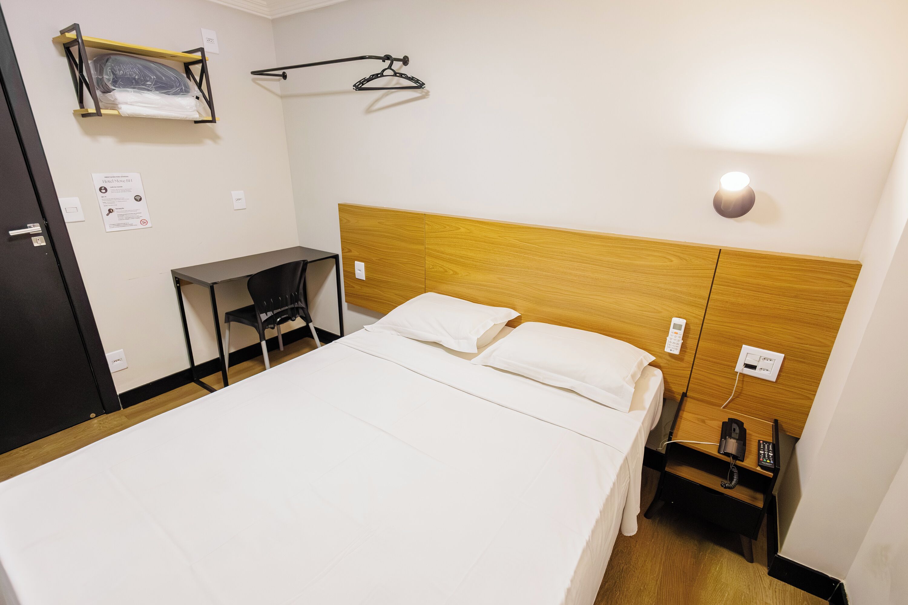 Habitación de hotel con cama, escritorio, silla, perchero y mesita de noche con teléfono.