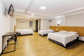 Ein Hotelzimmer mit drei Betten, einem Schreibtisch, einem Sessel, einem Fernseher und einer Klimaanlage.