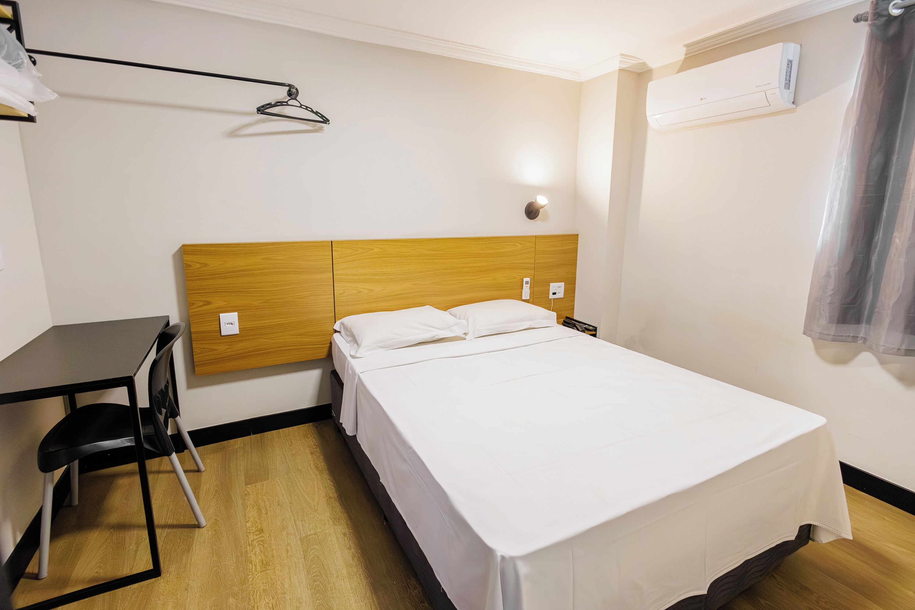 Ett hotellrum med en säng med ett huvudgavel av trä, ett skrivbord med en stol, en klädstång och en luftkonditioneringsanläggning.