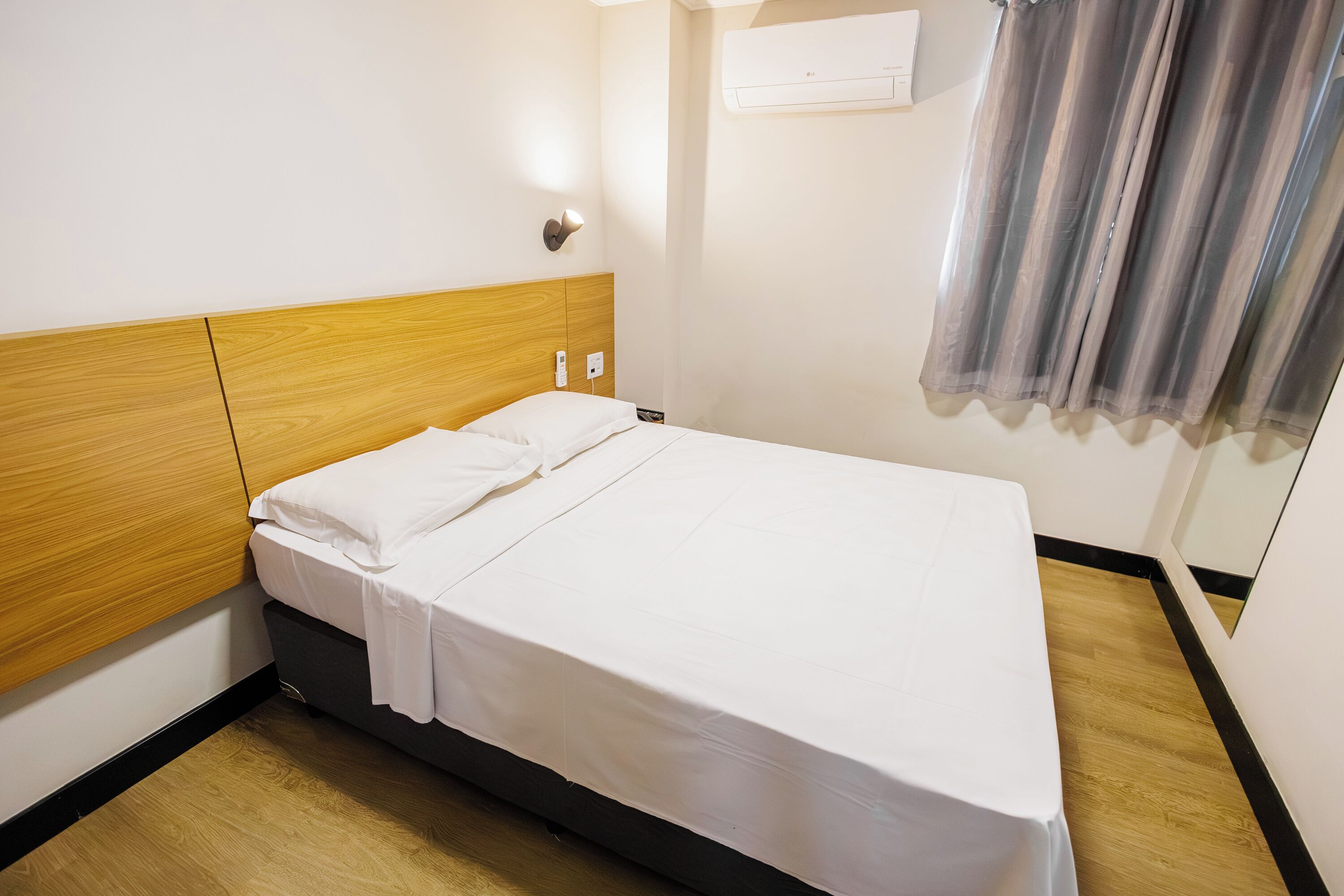 A hotel room with a wooden headboard, a bed with white linens, an air conditioning unit, and a curtain.