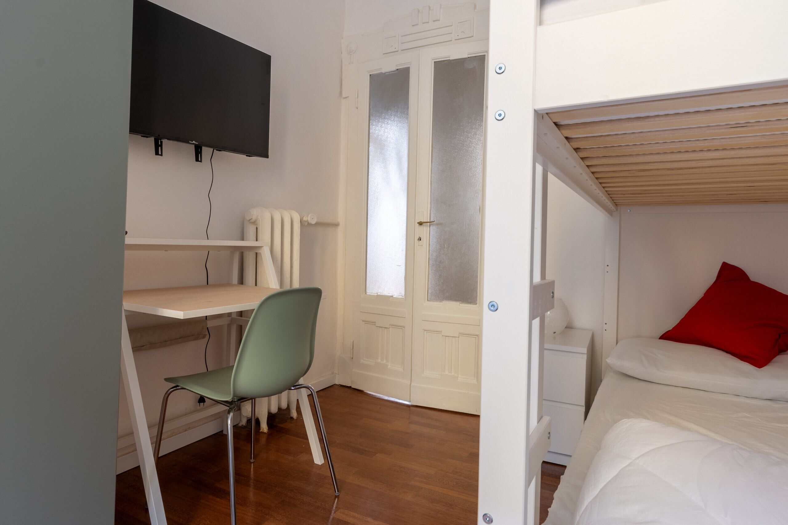 A room with a bunk bed, a desk with a chair, a television mounted on the wall, and a door with frosted glass panels.