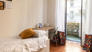 Une chambre à coucher comprenant un lit, un bureau, une chaise et une fenêtre donnant sur des immeubles.