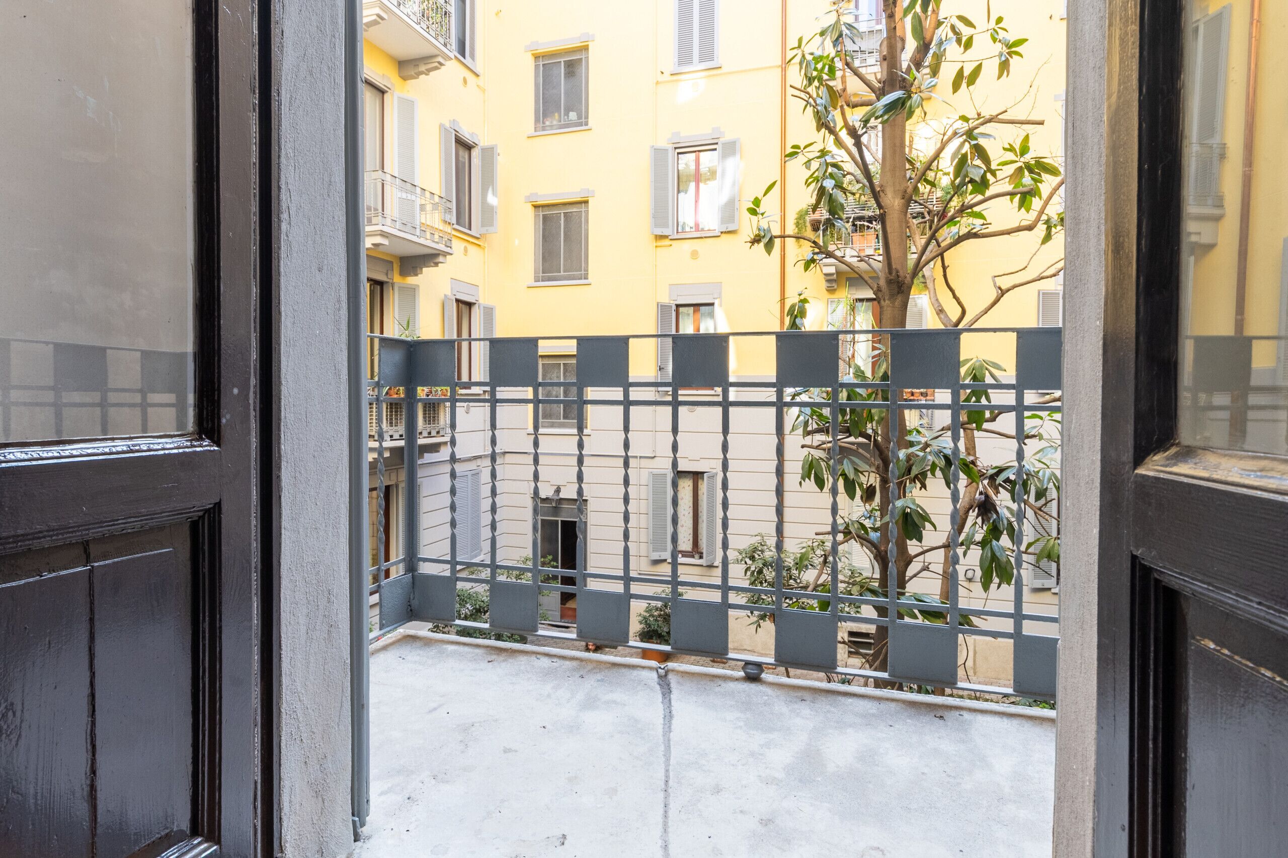 Un balcón con barandilla de metal, vista de un edificio amarillo con varias ventanas y un árbol en primer plano.