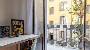 Une pièce avec une fenêtre donnant sur un balcon doté d’une balustrade en métal. On y trouve une chaise et une table sur laquelle sont posés un cadre avec une photo et une tasse.