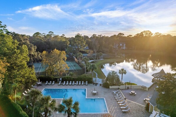 A resort with a swimming pool, sun loungers, a tennis court, and a lake view.