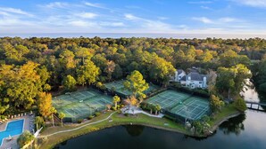 Eine Vogelperspektive auf einen Tennisplatzkomplex, umgeben von dichtem Wald, einem Schwimmbad und einem großen Haus.