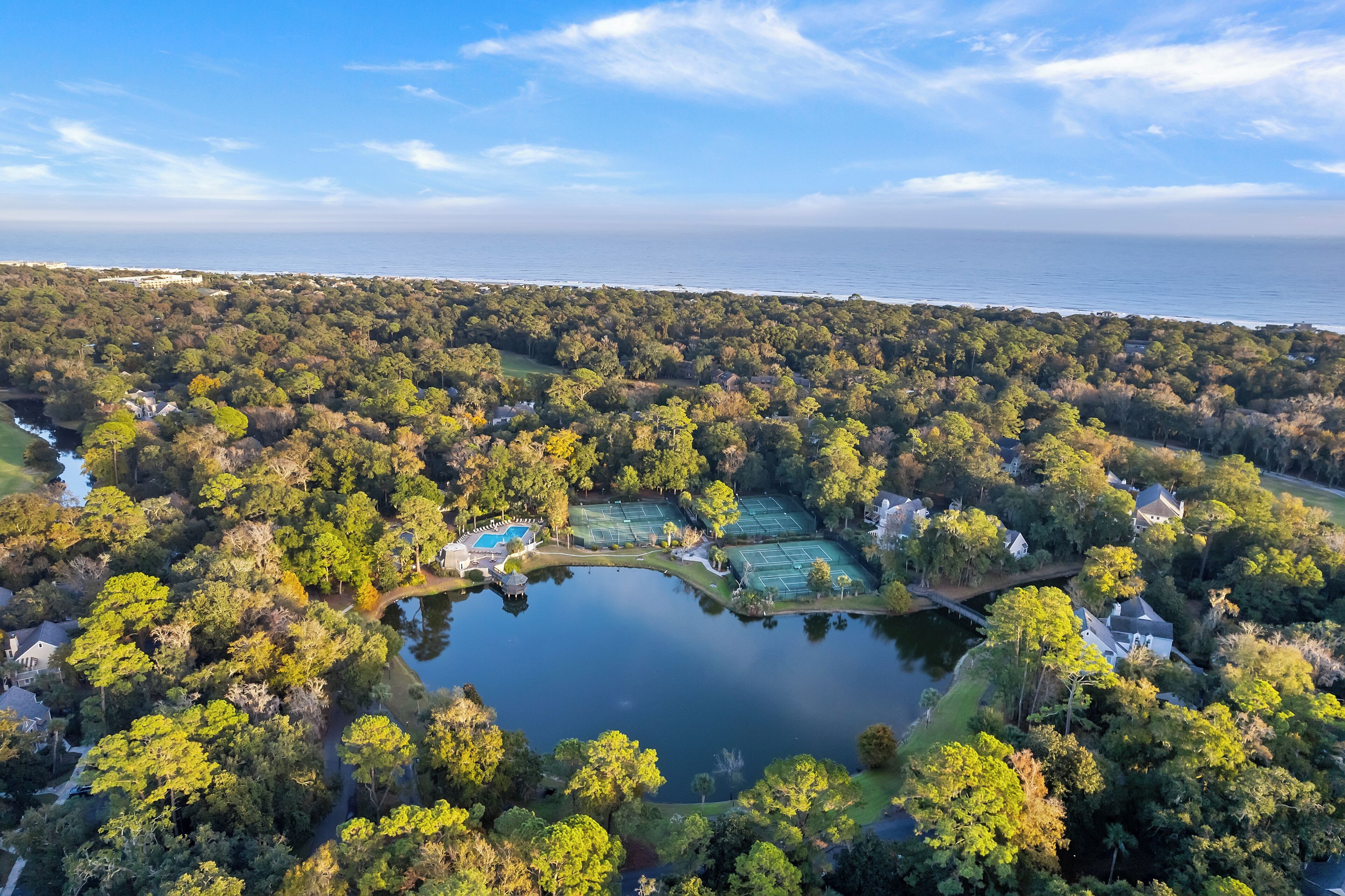 Vista aérea de una zona residencial con un lago, canchas de tenis y casas rodeadas de árboles frondosos.