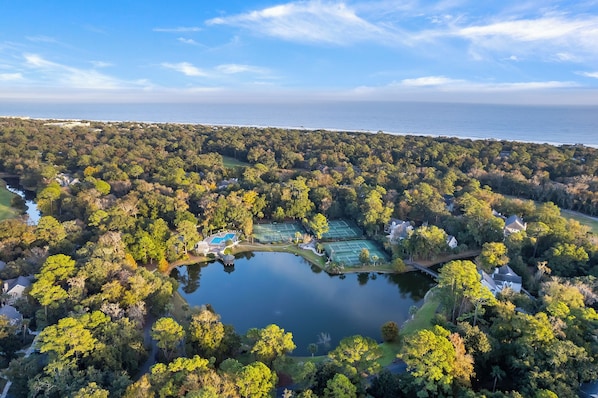 An aerial view of a residential area with a lake, tennis courts, and houses surrounded by dense trees.