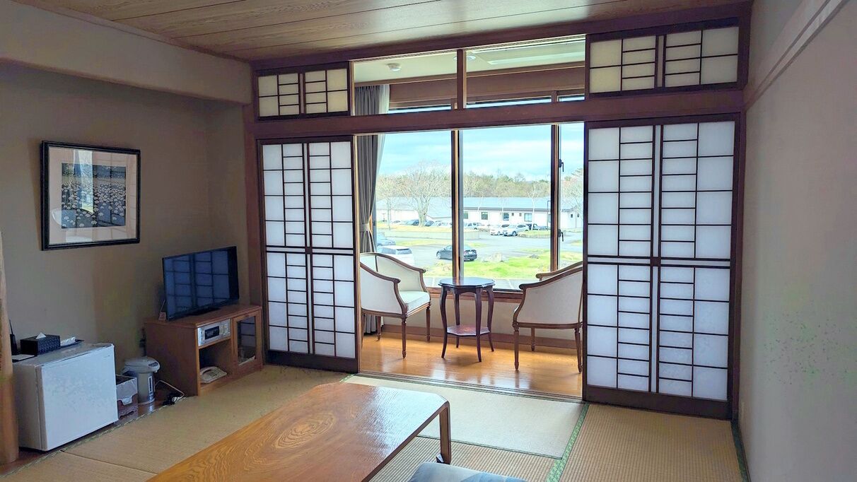 A traditional Japanese-style room with tatami flooring, a television, a small refrigerator, and a view of the outside.