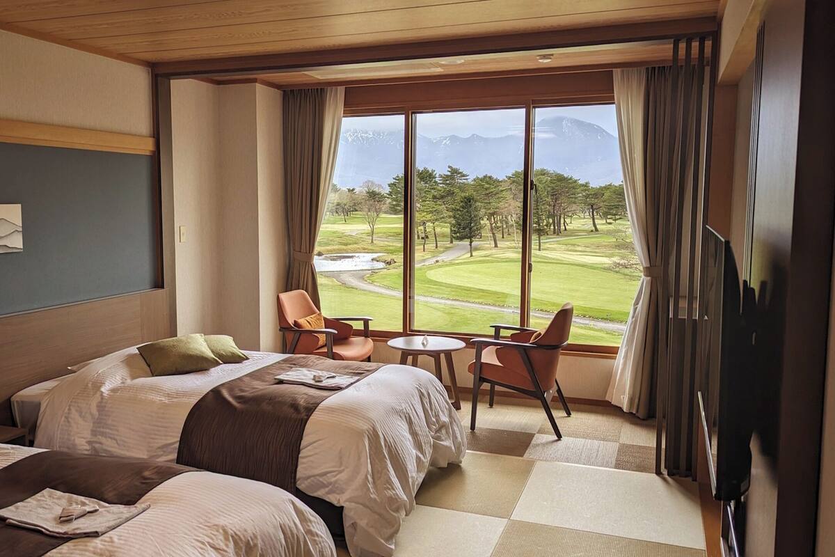 Une chambre d’hôtel avec deux lits, un coin repas comprenant une table et des chaises, et une grande fenêtre donnant sur un terrain de golf et des montagnes.