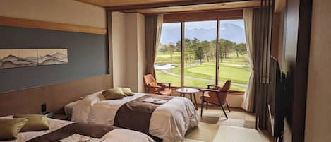 A hotel room with two beds, a dining area with a table and chairs, and a large window offering a view of a golf course and mountains.
