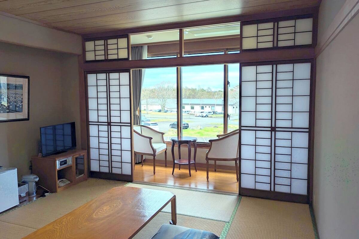 Une pièce de style japonais traditionnel, avec un sol recouvert de tatamis, une basse table en bois, un téléviseur posé sur un meuble et des portes coulissantes en papier de riz (shoji) donnant sur un balcon avec vue sur un parking.