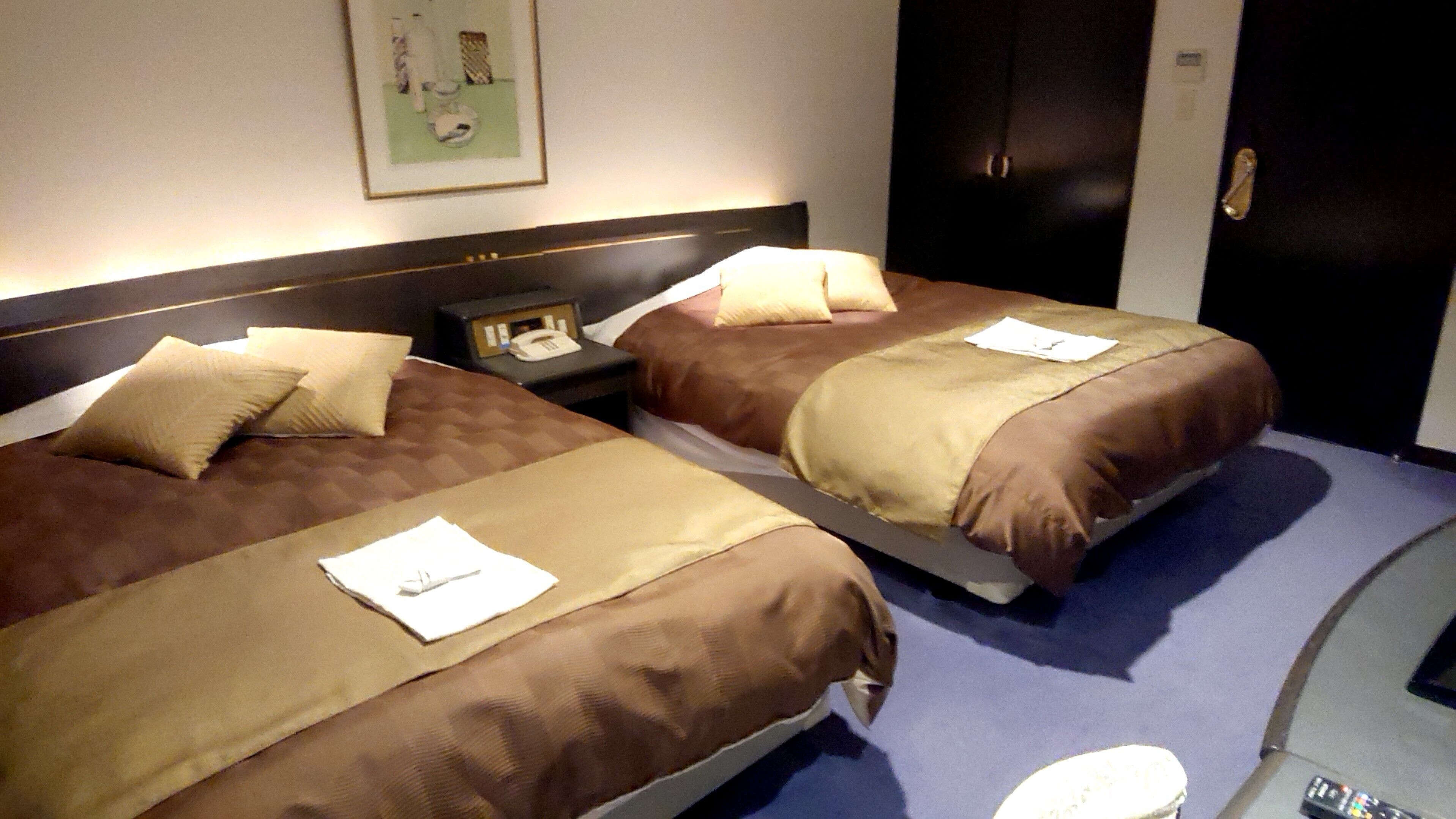 Dos camas con ropa de cama marrón, una mesita de noche con un teléfono y un cuadro en la pared.