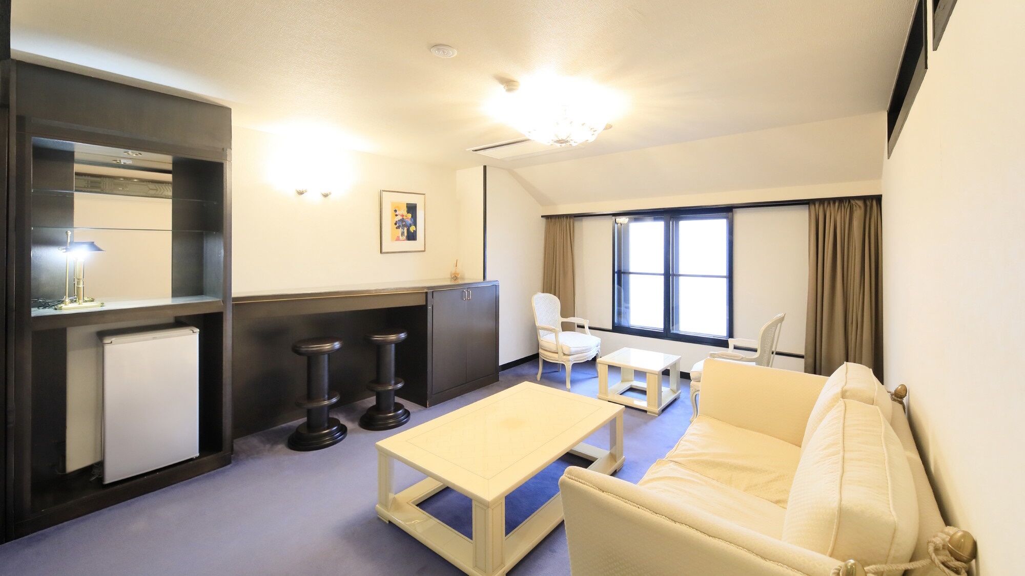 A compact hotel room with a sofa, a small table, a desk, a chair, a cabinet, and a window with curtains.