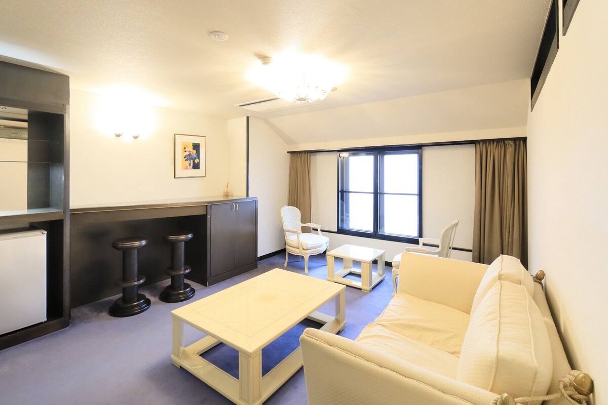Une chambre d’hôtel de taille réduite, comprenant un canapé, une petite table, un bureau, une chaise, une armoire et une fenêtre avec des rideaux.