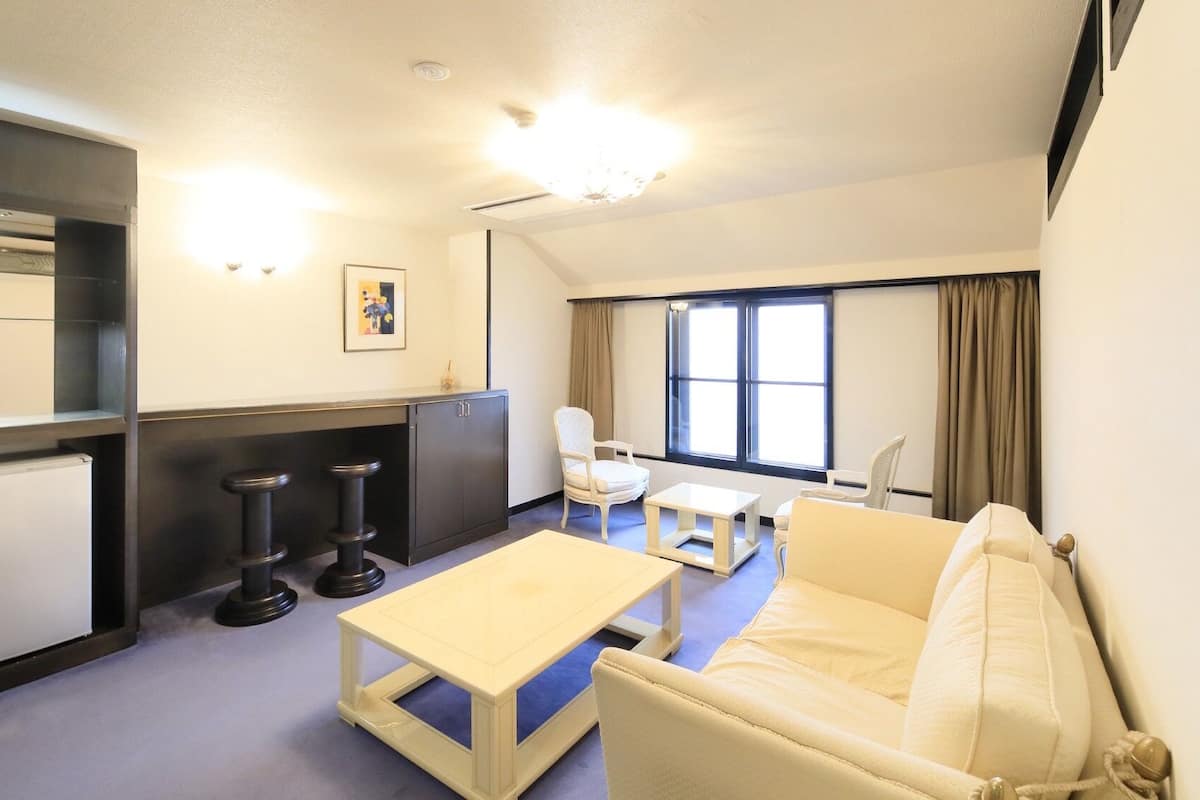 A compact hotel room with a sofa, a small table, a desk, a chair, a cabinet, and a window with curtains.
